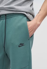 Χέρι στην τσέπη, φοράει τιρκουάζ παντελόνι φόρμας Nike με μαύρο κορδόνι και γκρι πουκάμισο, με ορατό τατουάζ στον πήχη.