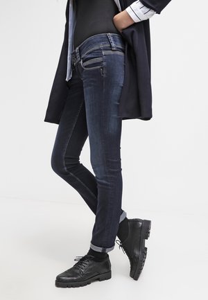 Donkerblauwe denim jeans met een strakke pasvorm, subtiele slijtage, ritssluiting en knoopsluiting, gecombineerd met zwarte gestructureerde veterschoenen.
