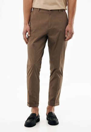 Mann trägt braune, eng zulaufende Hosen mit umgekrempelten Bündchen und schwarze Leder-Loafer, steht vor einem weißen Hintergrund.