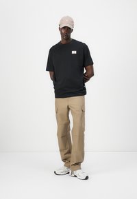 T-shirt noir à col rond avec un petit patch logo, associé à un pantalon cargo beige avec des poches, et des baskets blanches avec des accents noirs.