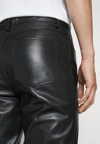 Svarta läderbyxor med en slät yta, med en distinkt bakficka och synliga sömnadsdetaljer. Midjan är smal och funktionell.