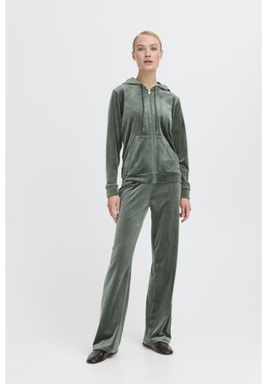 Zelen pulover z zadrgo iz velurja z žepi spredaj, kombiniran s primernimi dolgimi hlačami z ohlapnim krojem. Gladek otip in rahlo sijoč videz.