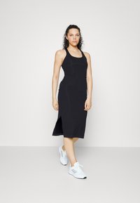 Vestido preto sem mangas com corpete ajustado, fenda lateral e comprimento médio. Modelo combinado com tênis brancos. Textura suave.