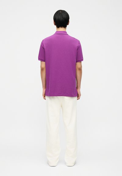 Άτομο που φοράει πορφυρό κοντομάνικο πόλο μπλουζάκι και άσπρο φαρδύ παντελόνι, στέκεται γυρισμένο πλάτη σε απλό λευκό φόντο.