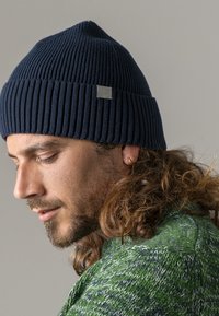 Navy ribbed knit beanie with a textured design and a small grey logo patch. Worn on a person with long, wavy hair and a green sweater.