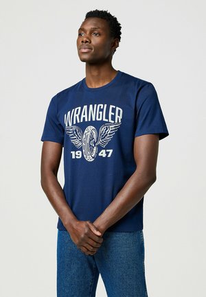 Mørkeblå kortærmet t-shirt med hvid grafisk print med vinger, "WRANGLER" og "1947" tekst. Lavet af bomuldsmateriale.