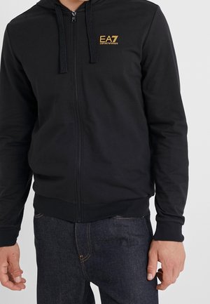 Czarna bluza z kapturem i zamkiem błyskawicznym z złotym logo "EA7 Emporio Armani" na piersi, noszona z ciemnoniebieskimi dżinsami, pokazana od szyi do bioder.