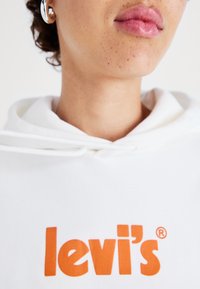 Vit hoodie med orange "levi's" logotyp. Har en dragsko i huvan och en mjuk textur som fokuserar på hals- och hakområdet.
