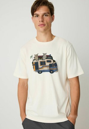 Jonge man met een wit T-shirt met een afbeelding van een camperbus en twee uilen erop, staand voor een effen achtergrond.