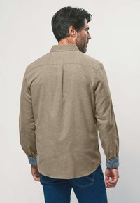 Mann mit dem Rücken zu sehen, der ein beiges Langarmshirt mit blauen Ärmelbündchen und dunkelblauen Jeans trägt, vor einem einfarbigen Hintergrund.