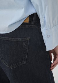 Ljusblå skjorta något instoppad i mörkblå jeans, där bakfickan och en del av en hand syns vid sidan.