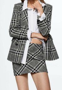 Blazer à carreaux noir et blanc avec une jupe mini assortie, présentant un tissu texturé, une coupe oversize et des revers classiques sur une chemise blanche.
