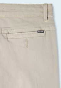 Un pantalon en coton beige présente une poche arrière avec une étiquette tissée, des détails subtils de couture et une texture lisse. La poche a un petit bord ourlé.