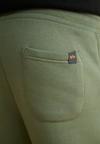 Oliven grønne sweatpants har et tekstureret stof, en baglomme med klap og et lille sort mærke med et gult logo syet på.