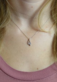 Zilveren ketting met een druppelvormige hanger met een heldere steen en fonkelende accentstenen aan één kant, gedragen tegen een roze top.