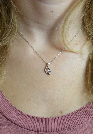 Zilveren ketting met een druppelvormige hanger met een heldere steen en fonkelende accentstenen aan één kant, gedragen tegen een roze top.