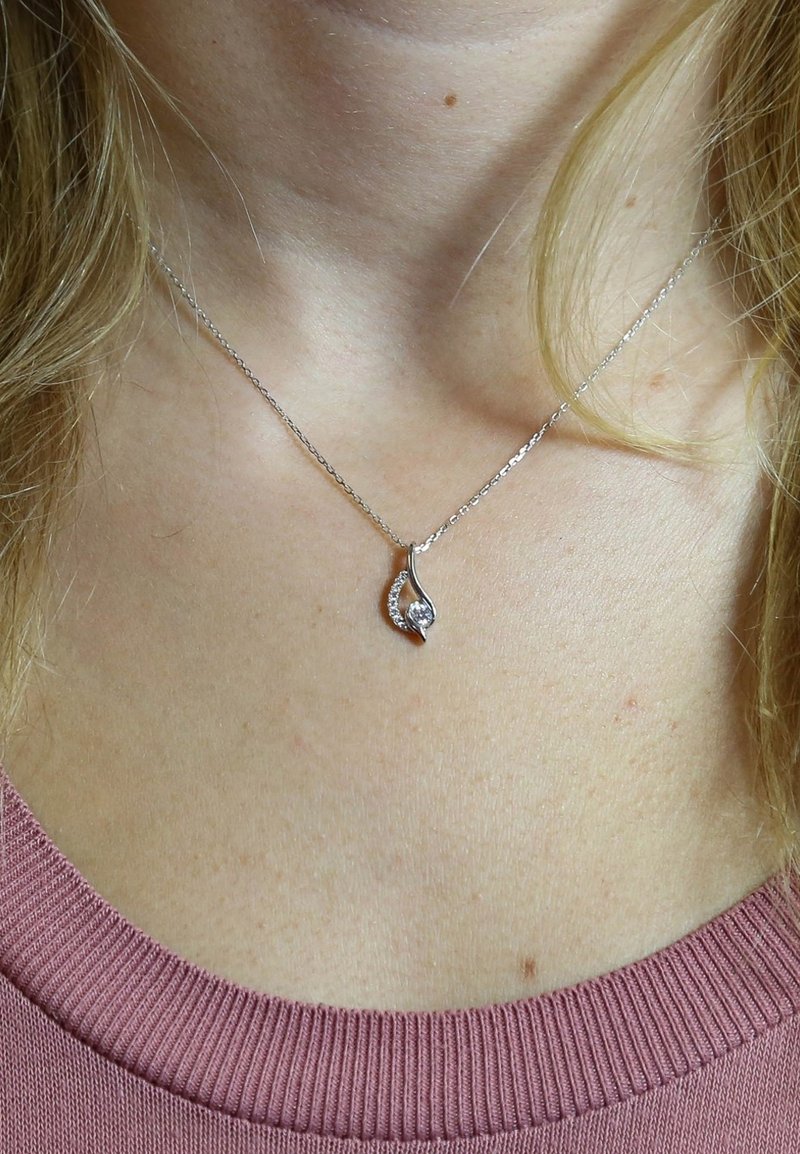 Zilveren ketting met een druppelvormige hanger met een heldere steen en fonkelende accentstenen aan één kant, gedragen tegen een roze top.