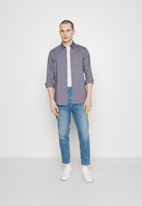 Bluza s gumbi v temno modri tkanini, dolgimi rokavi, zavihanimi navzgor. V kombinaciji z svetlo modrimi jeans hlačami in belimi supergami, stoji pred enobarvno ozadjem.