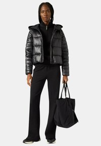 Giacca a vento nera con texture lucida e cappuccio, abbinata a una maglia scura a collo alto e pantaloni neri svasati, con una grande borsa tote nera in mano.