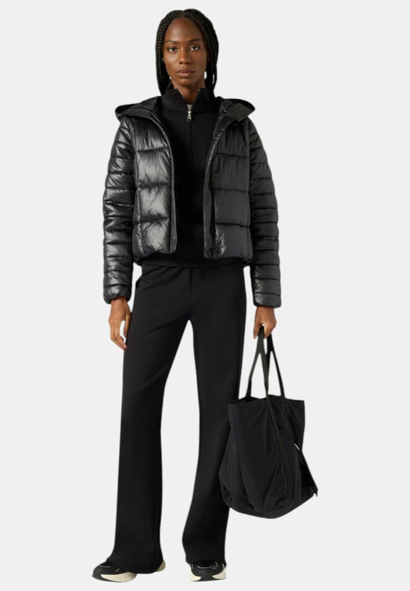 Giacca a vento nera con texture lucida e cappuccio, abbinata a una maglia scura a collo alto e pantaloni neri svasati, con una grande borsa tote nera in mano.