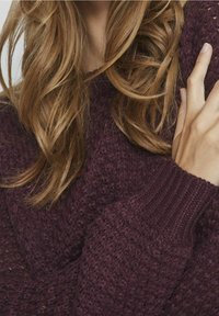 Burgundy texturerad stickad tröja med ribbad halsringning och manschetter. Fuzzy fibrer skapar ett mjukt utseende. Nära bild av tyg och handdetalj.