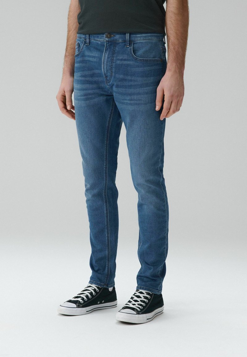 Mann trägt schmal geschnittene blaue Jeans und schwarze flache Sneaker, steht vor einem einfarbigen hellen Hintergrund.