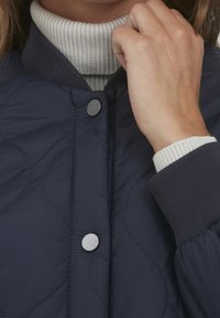 Käsi, joka säätää tummansinisen tikatun takin kaulusta kerma sävyisestä ribbikuvioidusta pooloneuleesta, näyttäen napit ja hihansuiden tekstiilin.