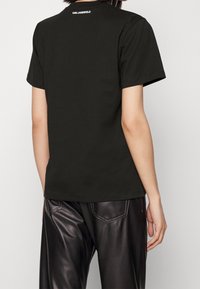 Camiseta de algodón negra de manga corta, con cuello redondo y un logo blanco en la parte trasera. Combinada con pantalones de cuero negros.