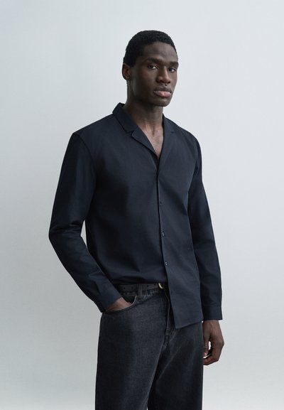 Homme à la peau foncée portant une chemise noire boutonnée et un jean foncé, debout devant un fond gris clair uni, une main dans la poche.