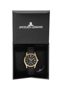 Guldtonad klocka med svart urtavla och läderrem, presenterad i en svart låda med en mjuk insida. Har datumfunktion och framträdande timmarkörer.