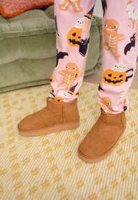 Braune Wildleder-Stiefeletten mit flacher Sohle, getragen zu rosa Pyjamahosen mit Halloween-Motiven, vor einem strukturierten Teppich.