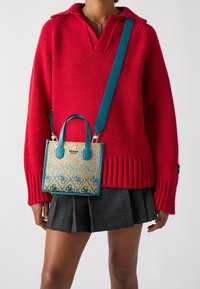 Rød strikket sweater med V-udskæring og ribbet kant. En lille, mønstret håndtaske lavet af vævet materiale med turkise detaljer hænger fra en strop.