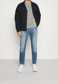 Schwarze Zip-Jacke über einem grauen T-Shirt mit Logo, kombiniert mit hellblauen geraden Jeans und weißen Sneakers. Glatte Stofftexturen.
