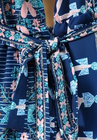 Námořnická modrá látka s geometrickými a květinovými vzory v teal, růžové a bílé, uvázaná do mašle kolem zipového uzávěru.