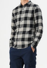 Zwart en grijs geblokt flanellen shirt met een kraag, lange mouwen en knoopmanchetten. Heeft een afgeronde zoom en donkere knopen.