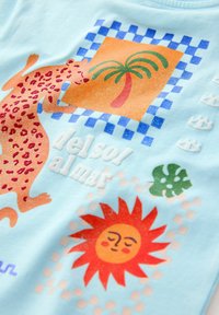 T-shirt bleu clair avec un graphisme d'un léopard, un palmier, un soleil et le texte "del sol al mar" dans des couleurs vives et des textures variées.