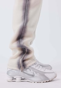 Gambe inferiori che indossano pantaloni beige chiaro con zip laterali sopra sneaker Nike bianche e argento con suole a cuscinetto d'aria visibile.