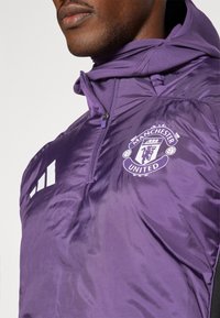 Lilla jakke med lynlås-krave, prydet med Manchester United-logo og hvide Adidas-striber på venstre side. Glat, skinnende stof.