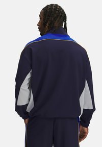 Chaqueta de chándal de color navy con cuello azul, paneles grises, ribetes amarillos y puños elásticos; detalles de costura visibles en los hombros y las mangas.