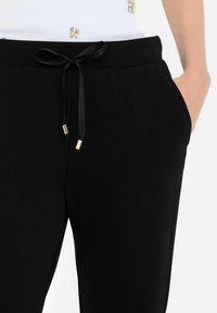 Črne tapered trenirke z vrvico v pasu z zlatimi detajli in stranskimi žepi, izdelane iz gladkega, lahkega materiala.