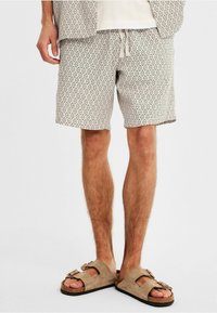 Shorts gris à motif en losanges, en tissu léger, avec un waistband élastique et un cordon de serrage. Assortis avec des sandales en suède beige.