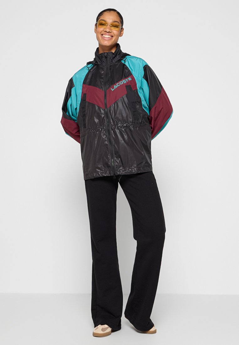 Czarna kurtka przeciwwiatrowa z akcentami w kolorze turkusowym i bordowym; zamek z przodu, regulowany dół i komfortowy krój. Połączona z czarnymi spodniami i trampkami.