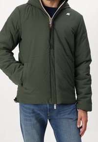 Mand iført en lynlås, mørkegrøn hættejakke med sidelommer og blå jeans, stående med den ene hånd i lommen.