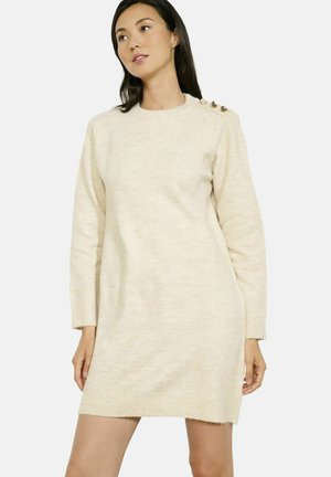 Donna che indossa un vestito maglione beige a maniche lunghe con dettagli di bottoni sulla spalla sinistra, in piedi davanti a uno sfondo bianco.