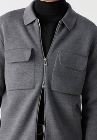 Šedá bunda na zip s límcem, dvěma náprsními kapsami, texturovanou látkou a hladkým zipem. Nošena přes černý rolák.