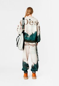 Conjunto de sudadera y pantalones extragrandes en beige, verde y blanco con patrones de montaña; presenta un logotipo destacado y puños elásticos; complementado con un bolso gris.