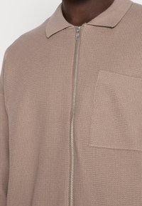 Primo piano di una camicia zip-up taupe a textured con colletto e taschino sul petto indossata da una persona con pelle scura.