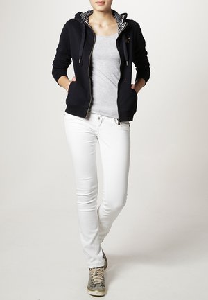 Felpa con zip blu navy con fodera interna a righe, canotta grigia, jeans skinny bianchi e sneakers grigie. La felpa presenta tasche frontali e coulisse.