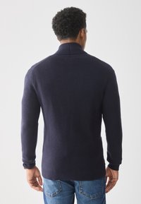 Granatowy prążkowany sweter z wysokim kołnierzem i długimi rękawami, o dopasowanym kroju zwężającym się ku talii. Gładka faktura.