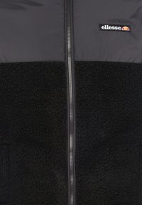 Sort zip-jakke med en glat overdel og en tekstureret, blød fleece-nederdel. Har et broderet logo på brystet.